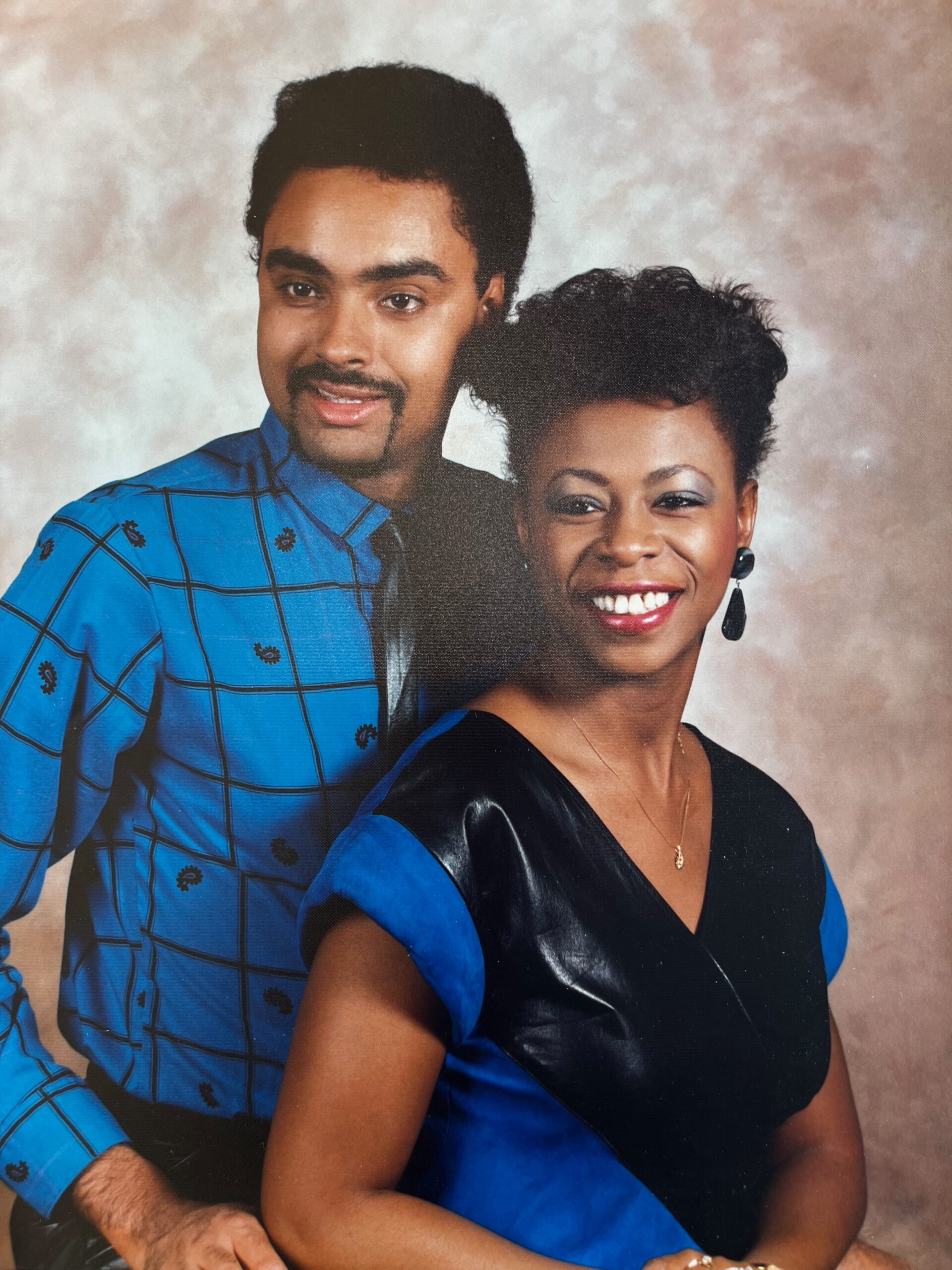 Smiling couple in a studio portrait; man in a blue plaid shirt behind a woman in a black and blue dress.