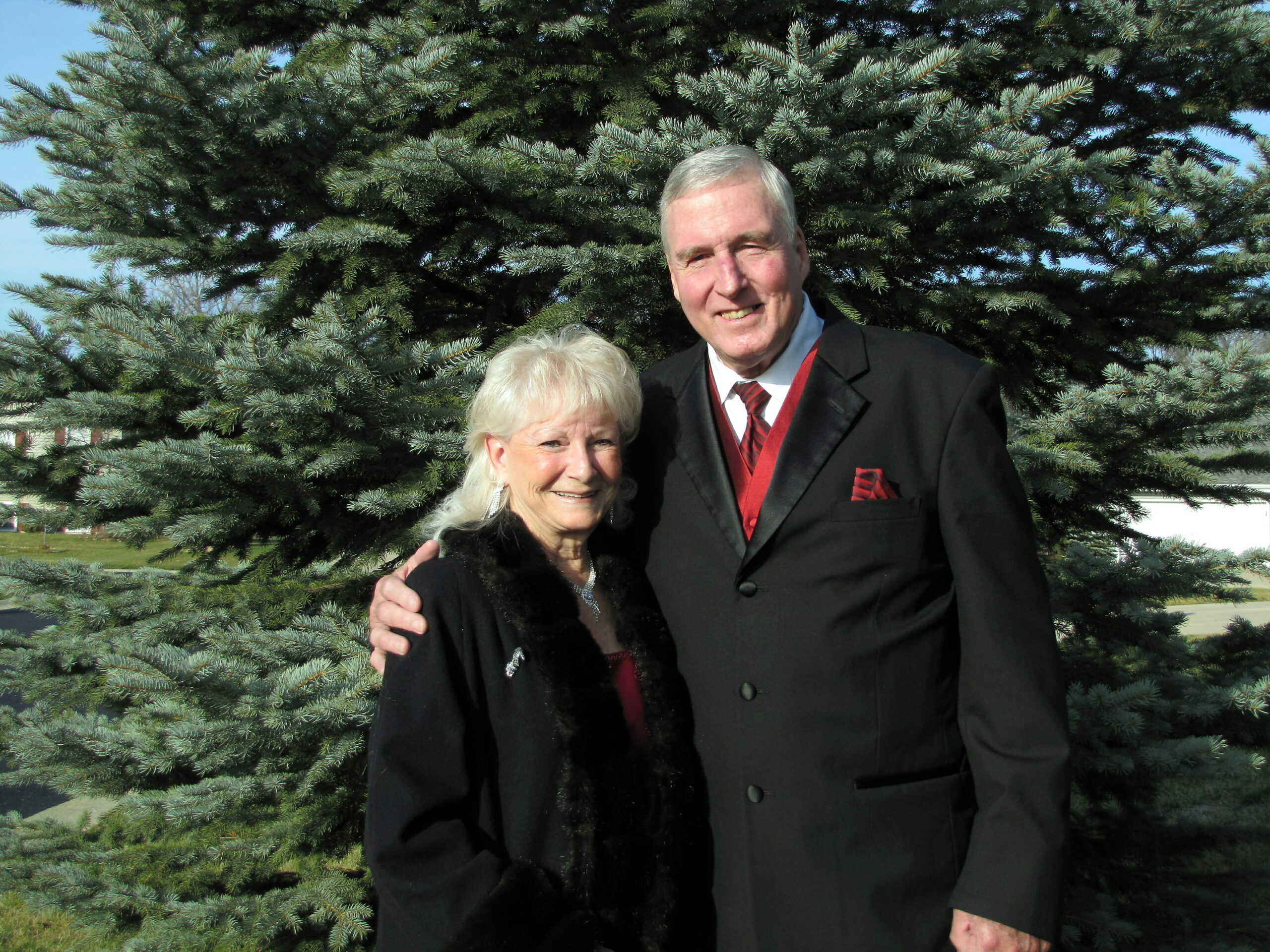 Smiling elderly couple in formal black coats posing in front of a large evergreen tree outdoors.