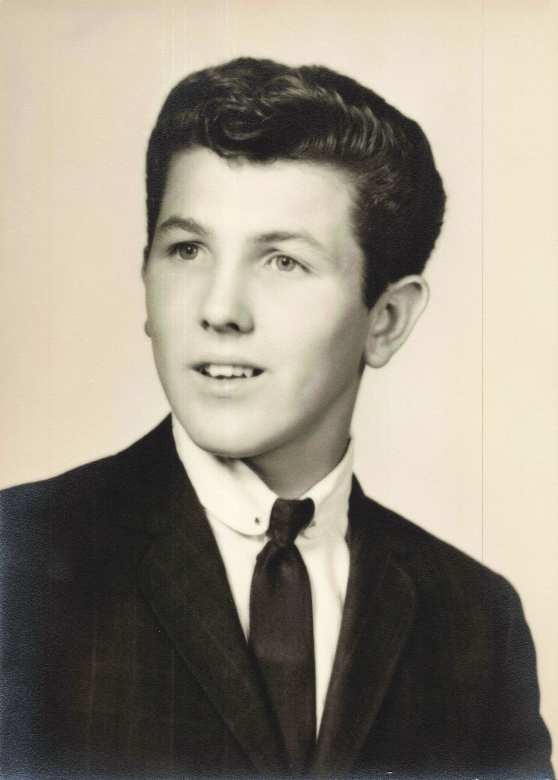 Black-and-white portrait of a young man in a suit and tie, looking slightly to the right with a faint smile across the lips.