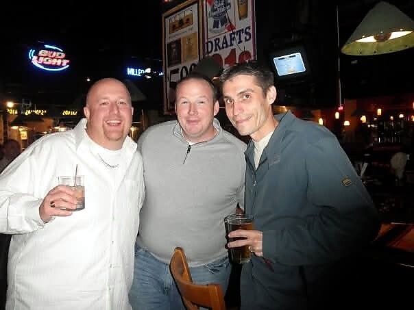 Three men posing together at a bar, smiling, each holding a drink, enjoying a night out.