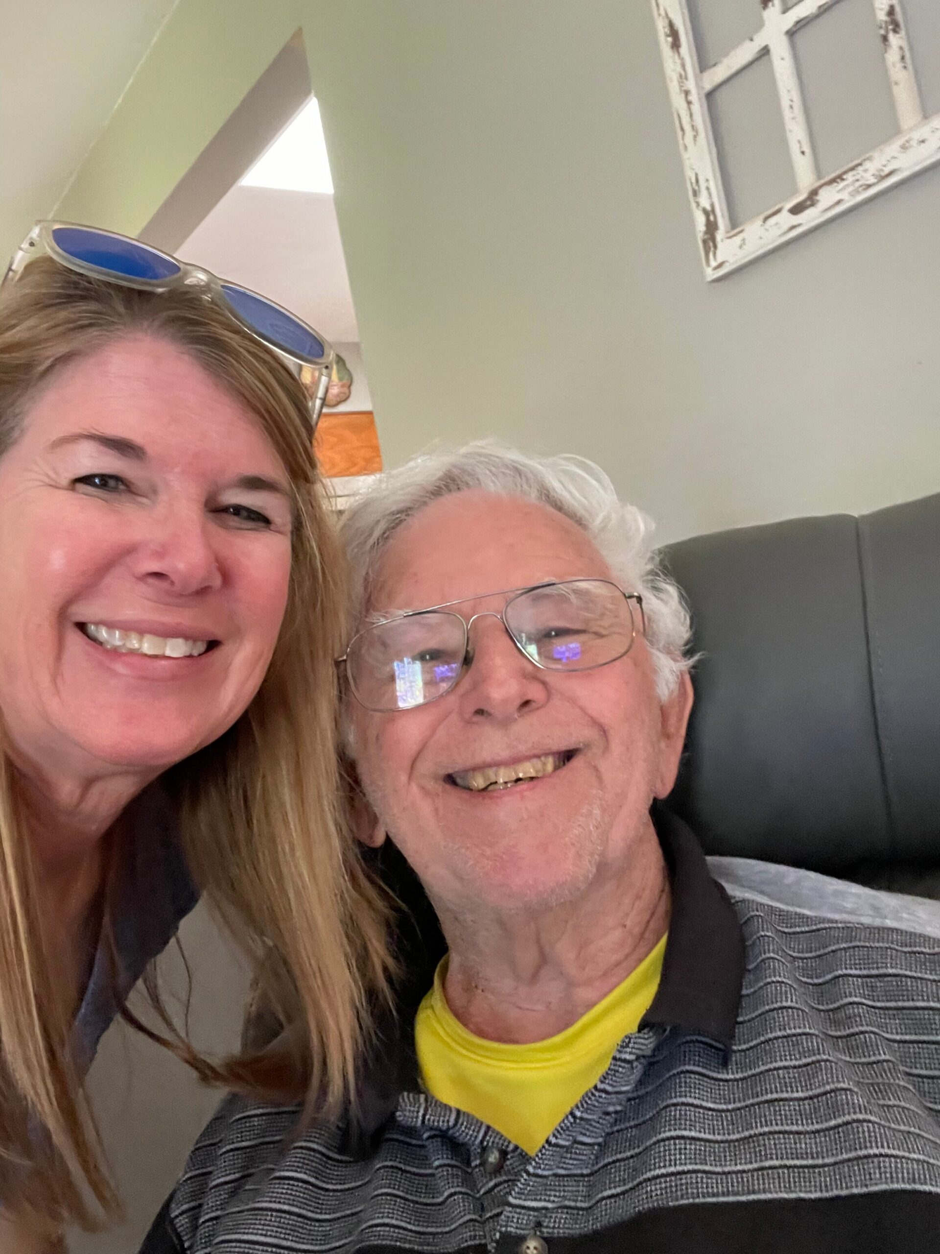 Close-up selfie of two people smiling indoors: a woman with sunglasses on her head next to an older man wearing glasses.