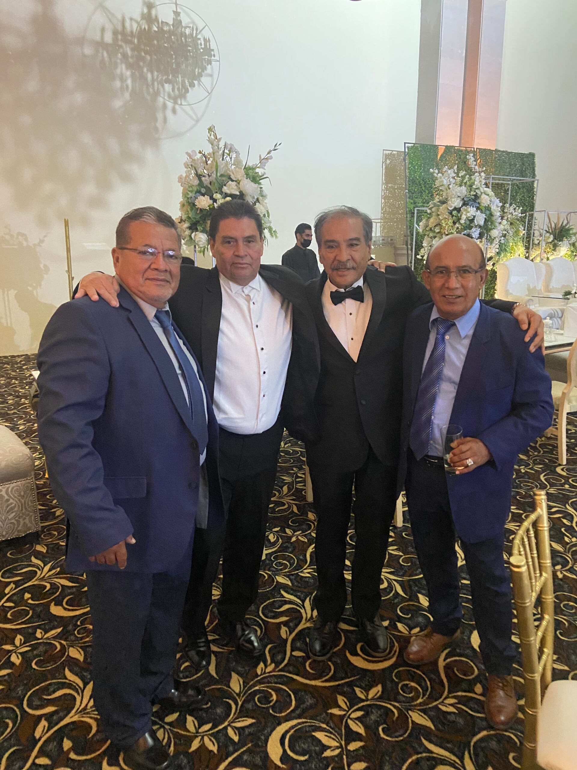 Four men in suits posing together at a formal event, with floral arrangements and decorative backdrop in a banquet hall.