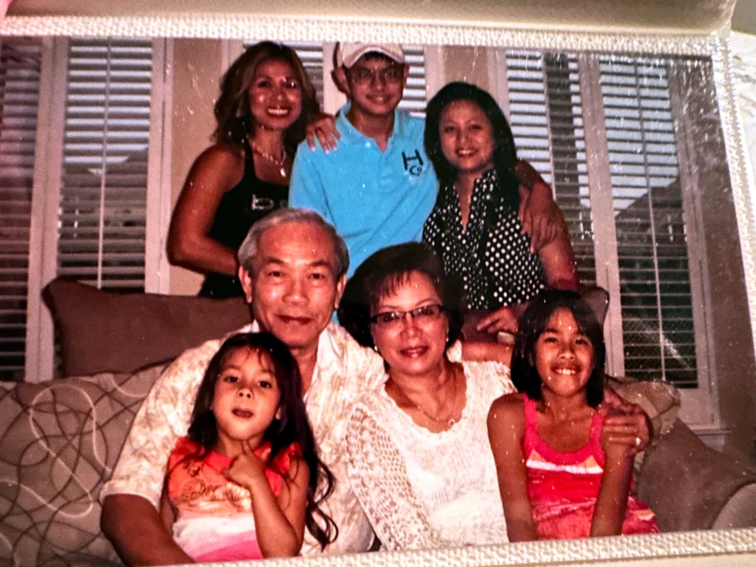 Smiling family of seven posing on a couch indoors with blinds behind them.
