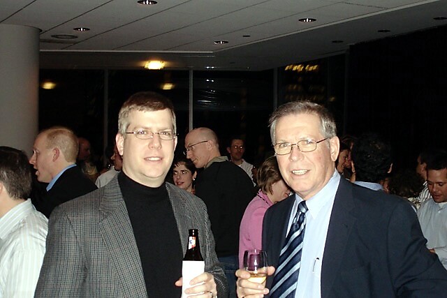 Two men in business attire at a crowded indoor reception, each holding a drink (one bottle, one glass) and smiling at the camera.