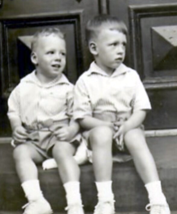 Two young boys sit side by side on steps in front of a door, wearing matching light shirts and socks, looking off to the side.