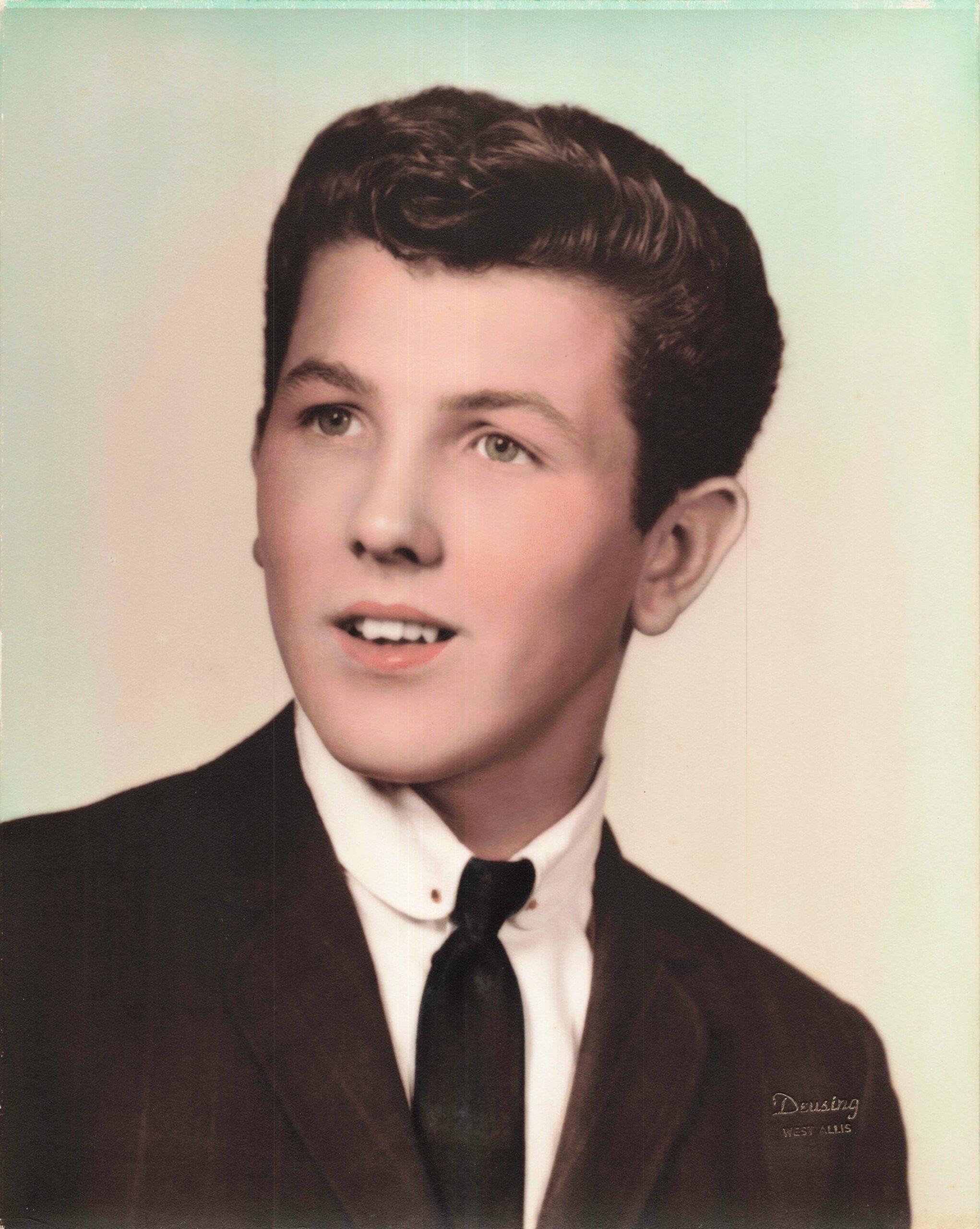 Youthful man in a dark suit and tie posing for a formal portrait in a studio setting, head and shoulders visible.