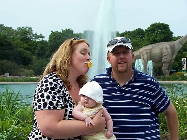 A woman holds a baby while a man in a striped polo stands beside them near a fountain and a dinosaur statue in a park setting.
