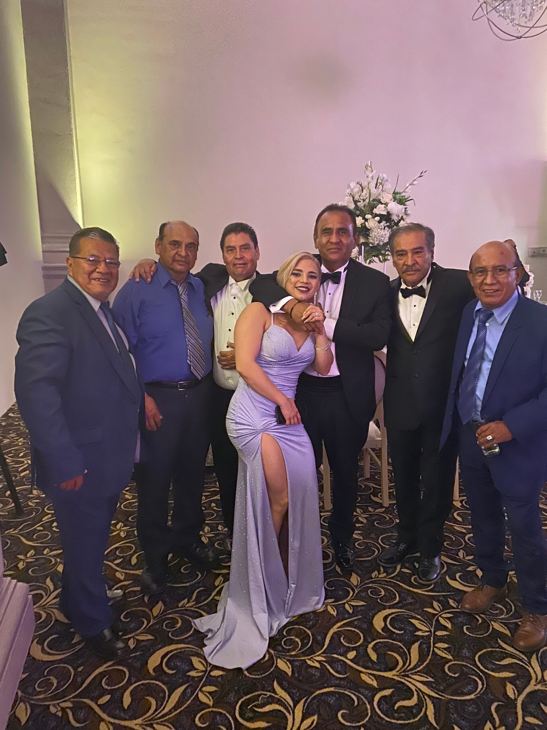 Group of eight adults in formal attire posing together at a banquet, woman in a lavender evening gown surrounded by men in suits and tuxedos.