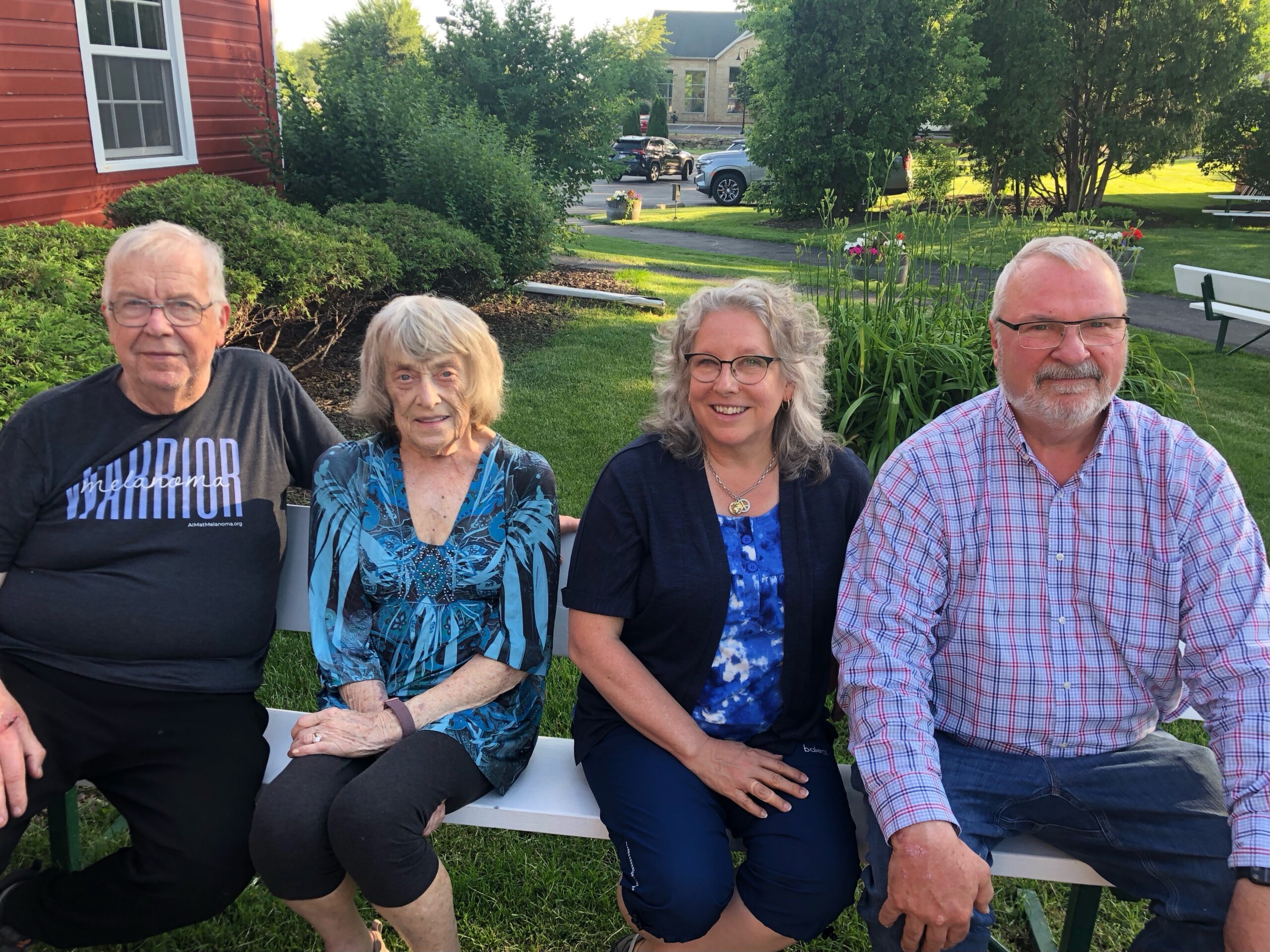 Four adults sitting on a white park bench outdoors, smiling, with greenery and a red building behind them.