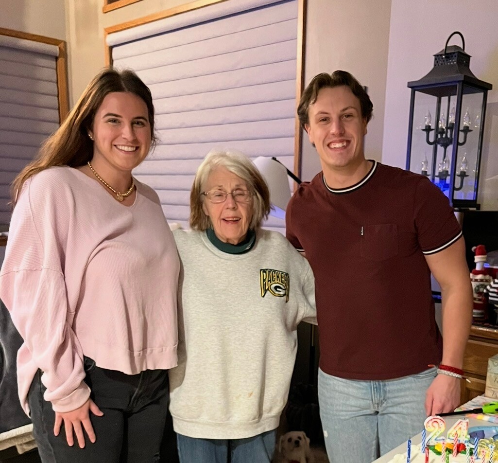Three generations posing for a birthday photo: a smiling young woman in a pink sweater, an elderly woman in a gray sweatshirt, and a smiling young man in a maroon shirt, with a birthday cake and candles in the foreground and a small dog on the floor behind them.