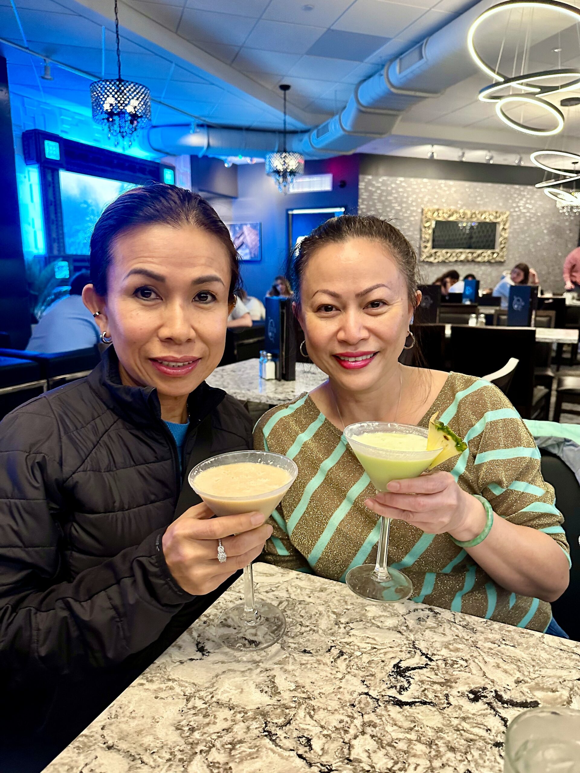 Two women sit at a marble table in a chic restaurant, toasting with pineapple-garnished cocktails.