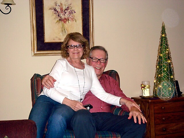 Couple sitting close together on a striped sofa in a living room, smiling at the camera with a festive tree on a dresser nearby.
