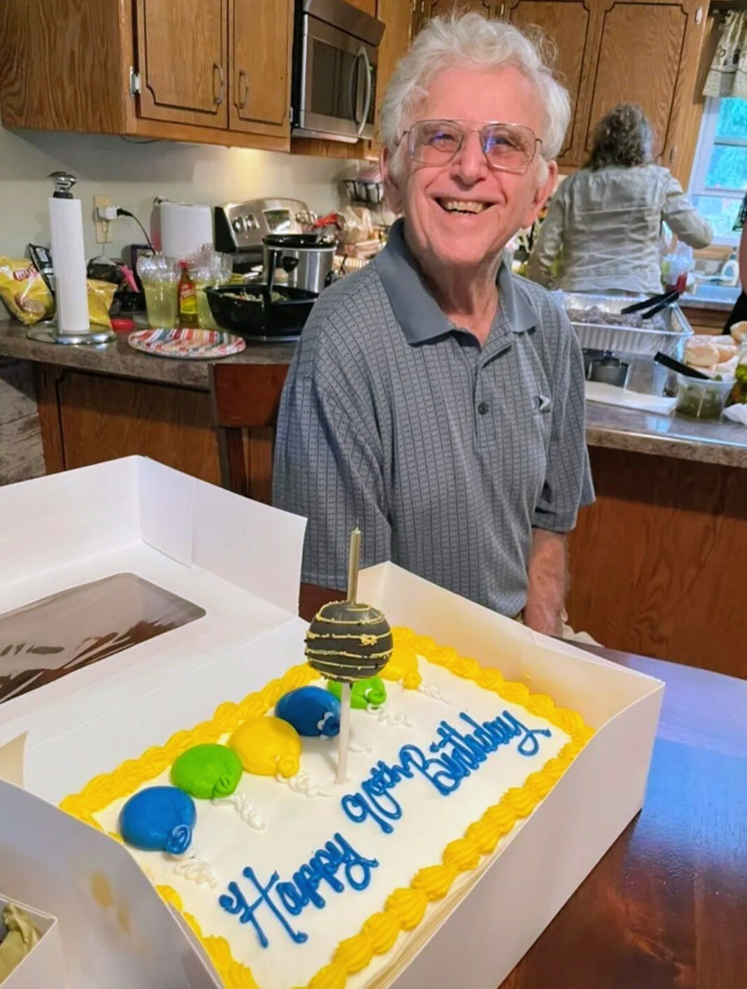 Smiling elderly man with white hair and glasses in a kitchen beside a birthday cake on the table.