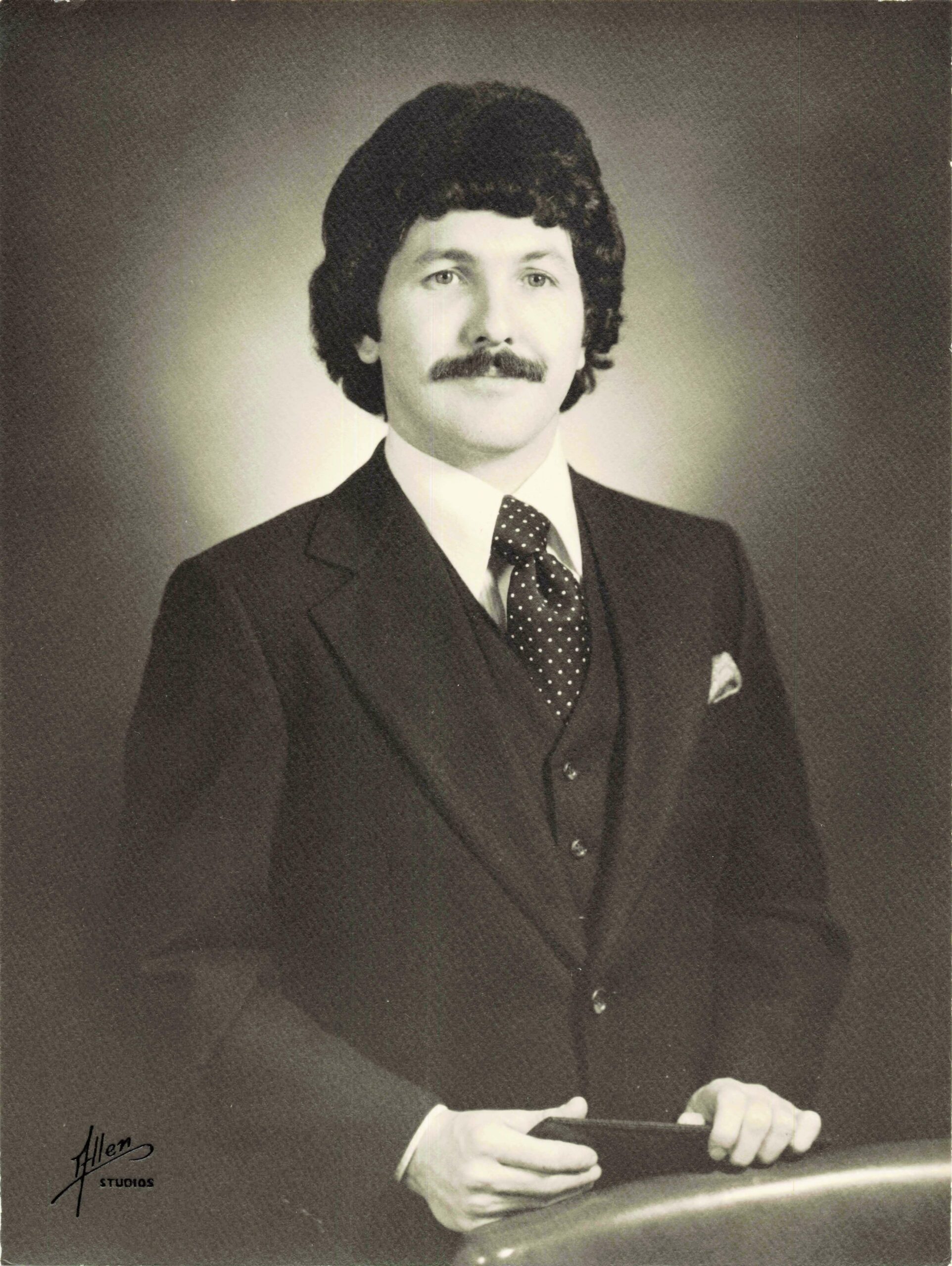Black-and-white formal portrait of a man with dark hair and mustache, wearing a suit, white shirt, and polka-dot tie, holding an item in his hands on a railing.