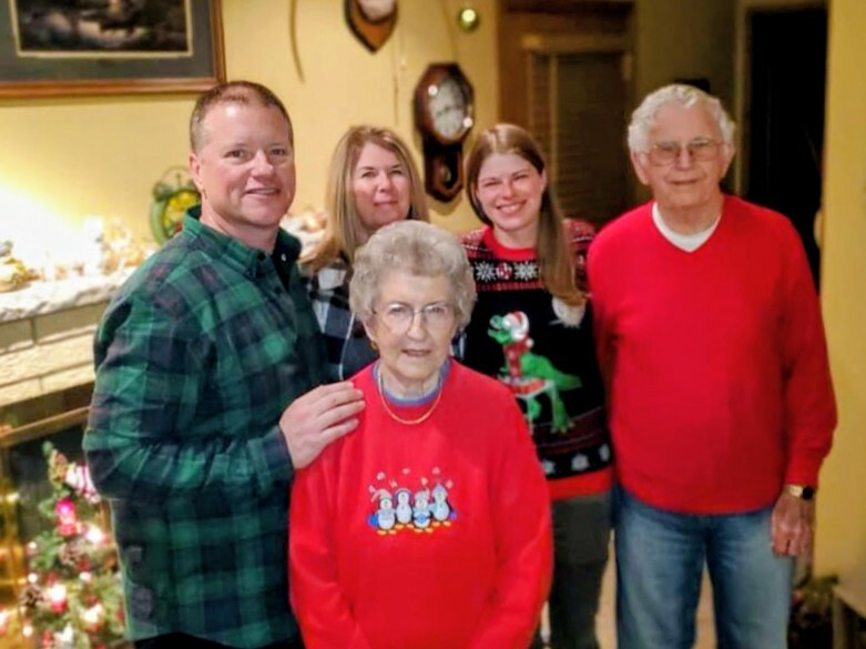 Five family members gather indoors for a Christmas photo, with an elderly woman in a red sweater front and center.