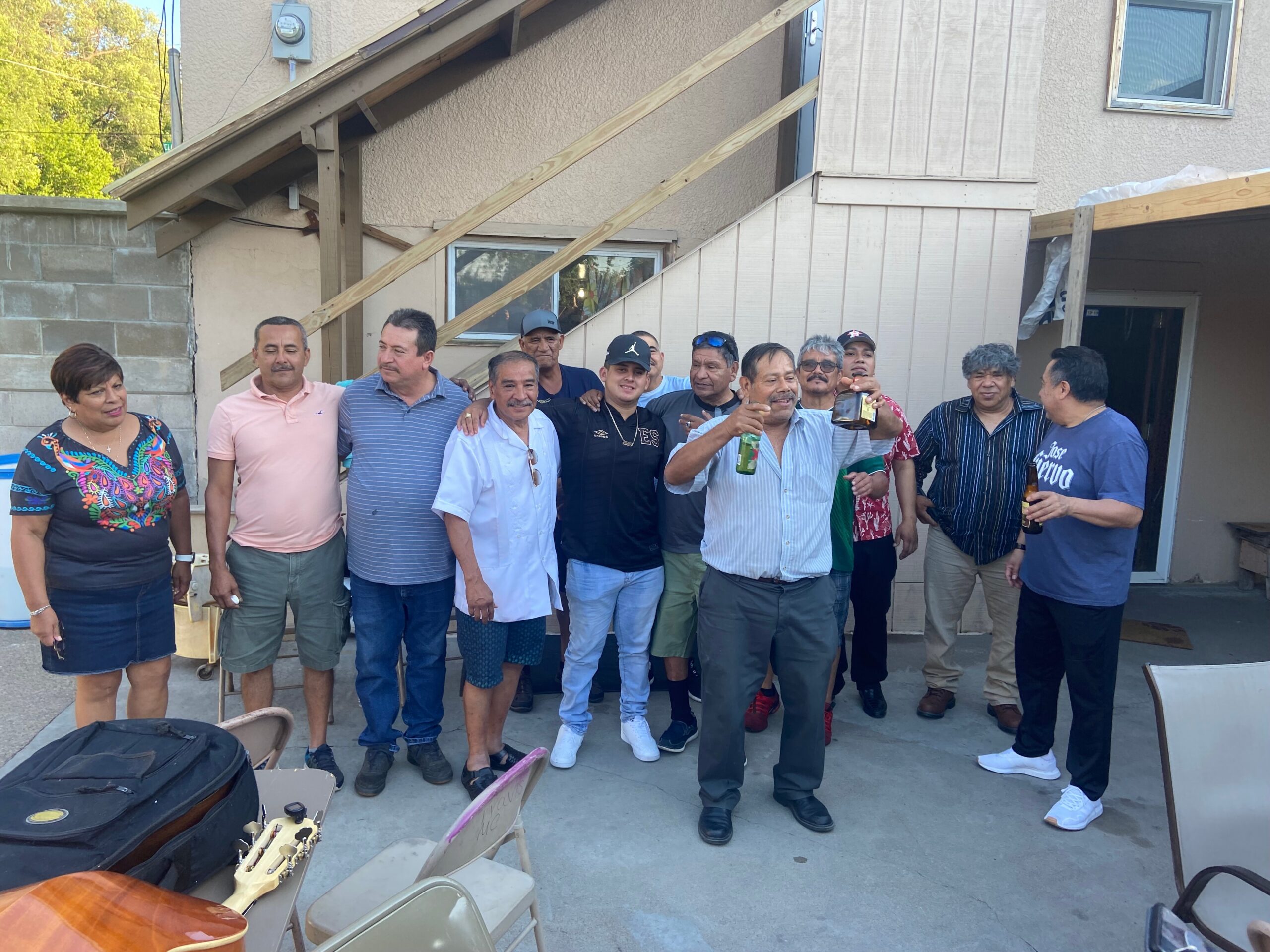 Group of adults posing together in a backyard or patio area, with guitars and chairs in the foreground and a beige building behind them.