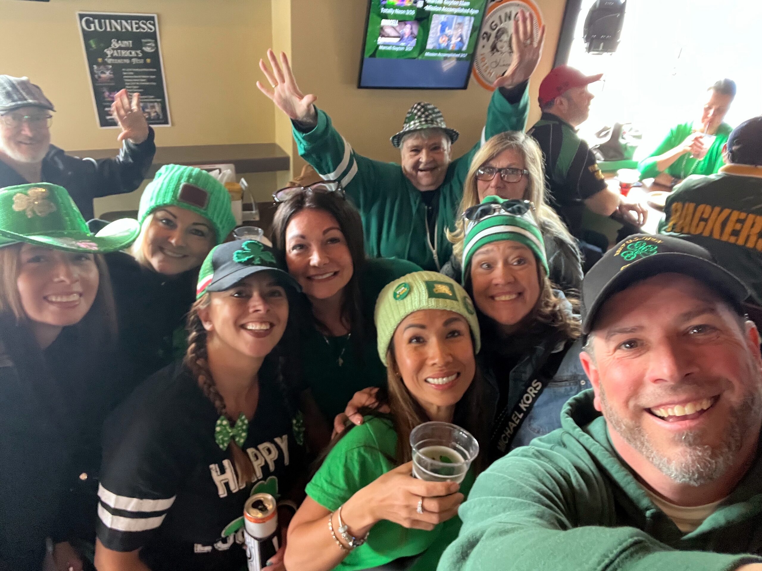 Group of friends dressed in green celebrating St. Patrick's Day, posing for a selfie in a bar with drinks and smiles.