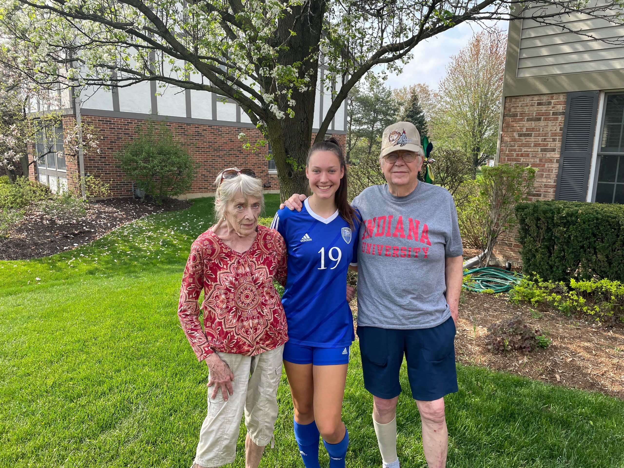 Three people pose together outdoors in a lawn yard with a flowering tree behind: grandmother, a smiling young athlete in blue sportswear, and grandfather wearing an Indiana University shirt and cap.