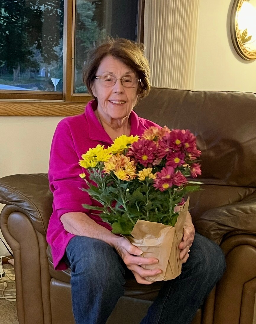 Older woman in a bright pink top sitting on a brown leather chair, holding a wrapped bouquet of yellow and pink mums.