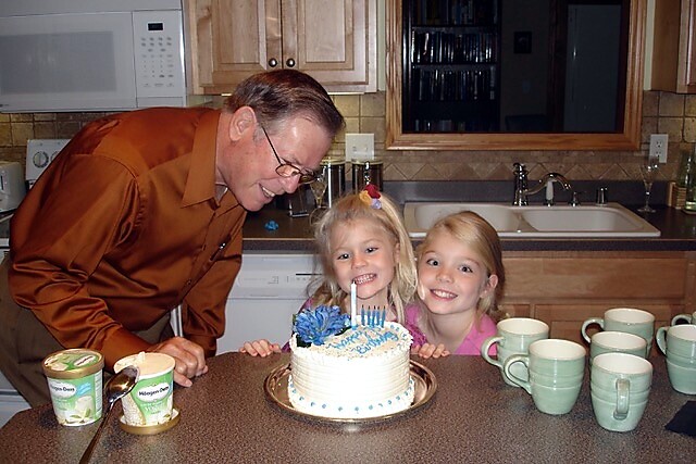 Man and two young girls lean in toward a frosted birthday cake with lit candles on a kitchen counter, smiling at the camera/each other.