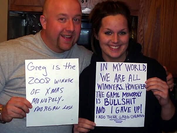 Smiling man and woman hold handwritten signs about Greg and Monopoly, posing for the camera.