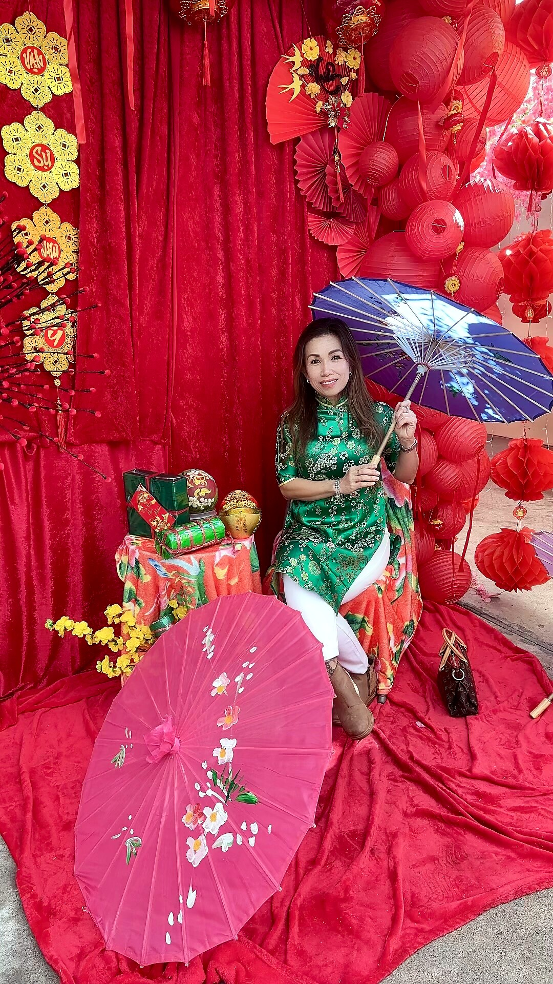 Woman in a green floral áo dài sits among red New Year decorations, holding a blue umbrella with a red backdrop and lanterns.