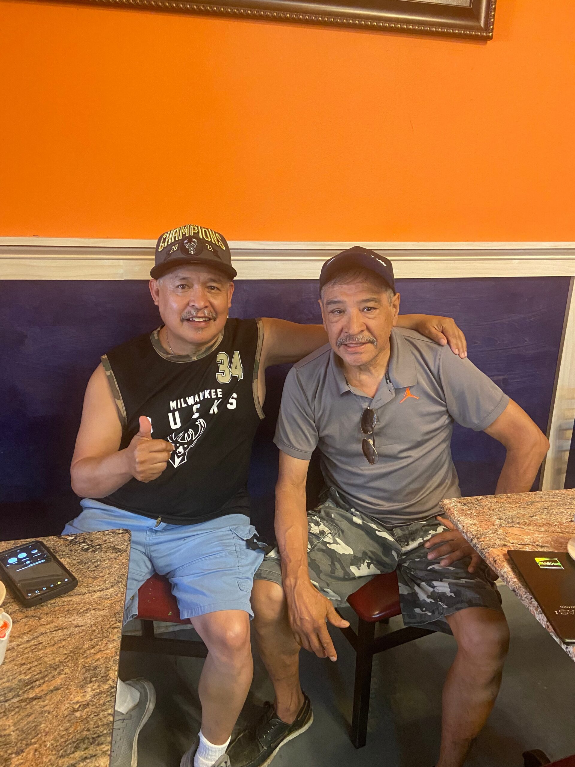 Two men sit at a counter in a restaurant, one giving a thumbs-up with a Milwaukee Bucks shirt and a cap that says Champions 2021, orange wall behind them.