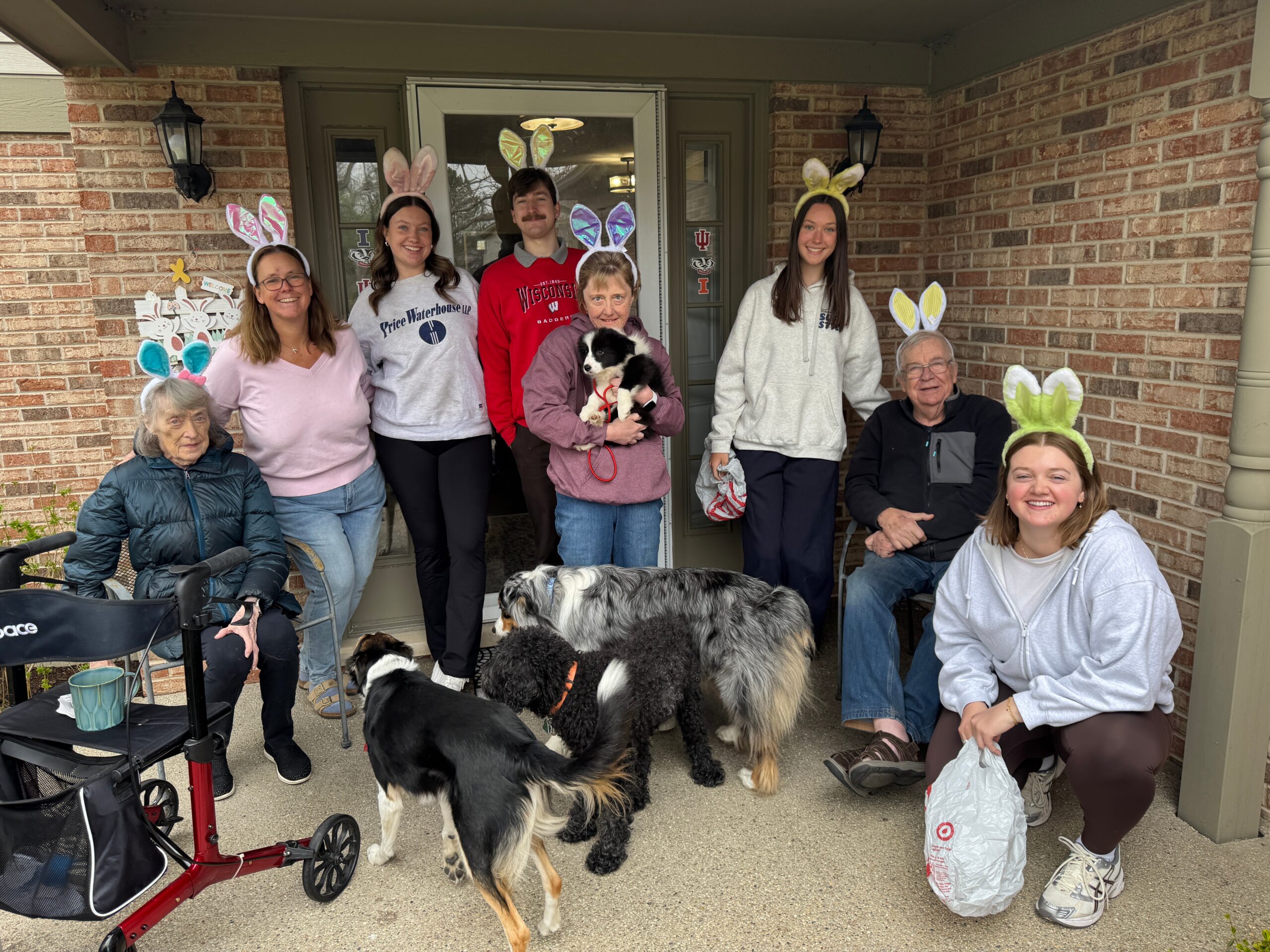 Group of people wearing Easter bunny ears pose on a brick porch with several dogs nearby.