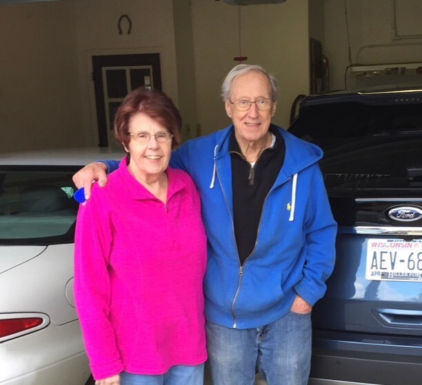 Elderly couple standing in a garage, arm in arm and smiling at the camera near parked cars.