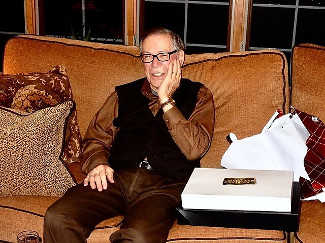 Older man with glasses sits on a tan couch, hand resting on his cheek, smiling, with a white gift box on a table beside him.