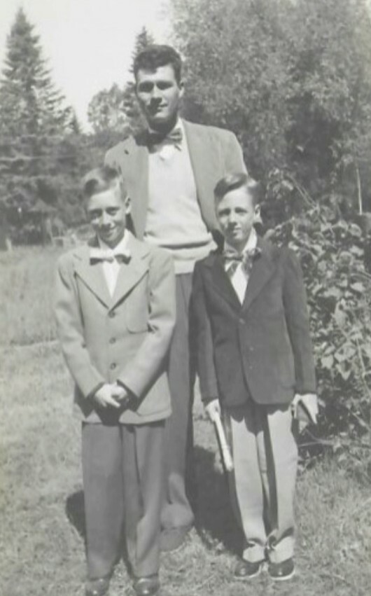 Three males posing outdoors in vintage-style suits: two young boys in bowties stand in front of an older man in a suit.