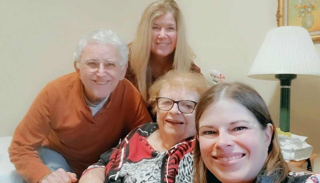Four people pose for a casual living-room selfie: an older man in orange, an elderly woman with glasses, a younger woman in front, and a blonde woman behind them, all smiling