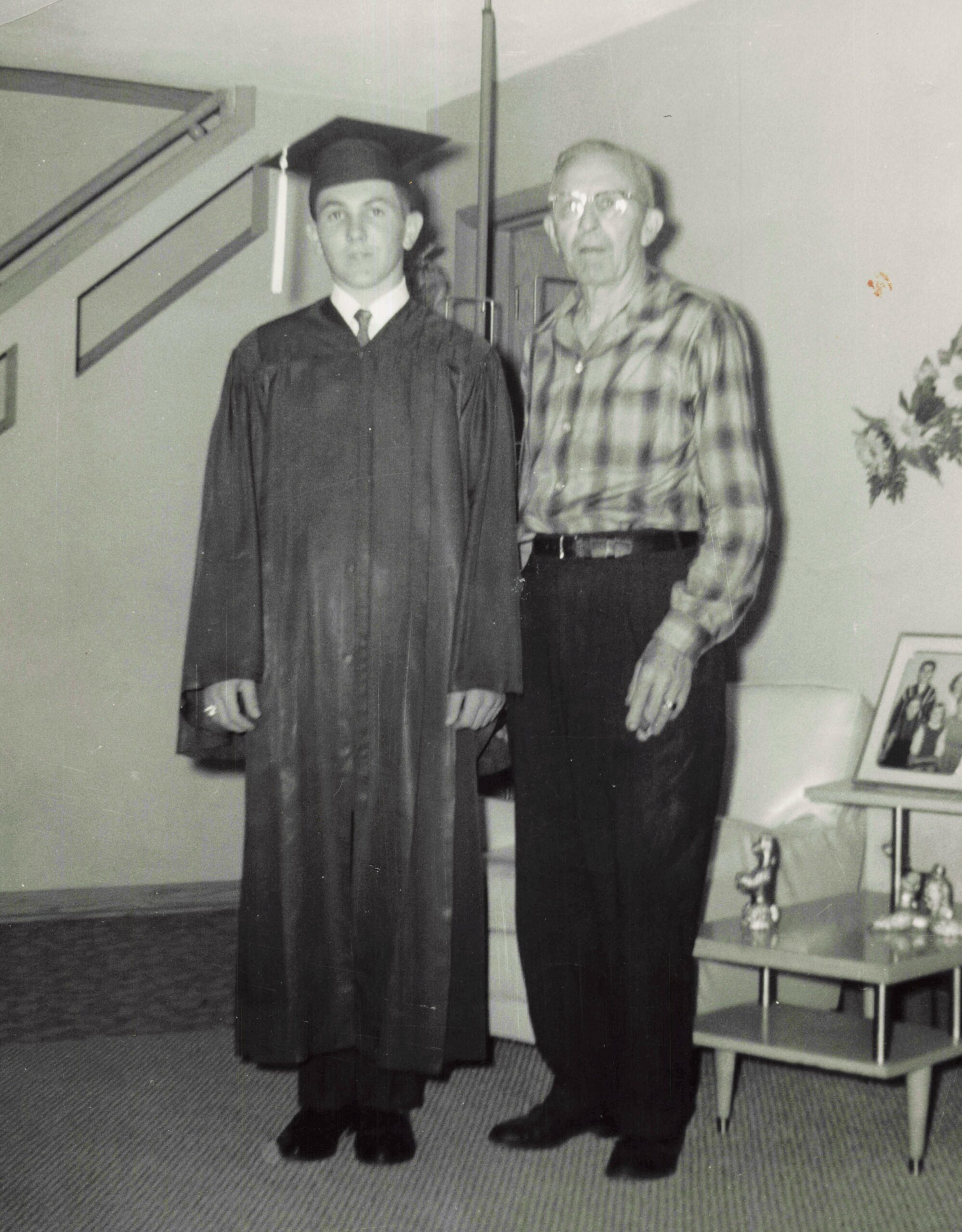 Young graduate in cap and gown posing indoors with an older man in a plaid shirt beside him.