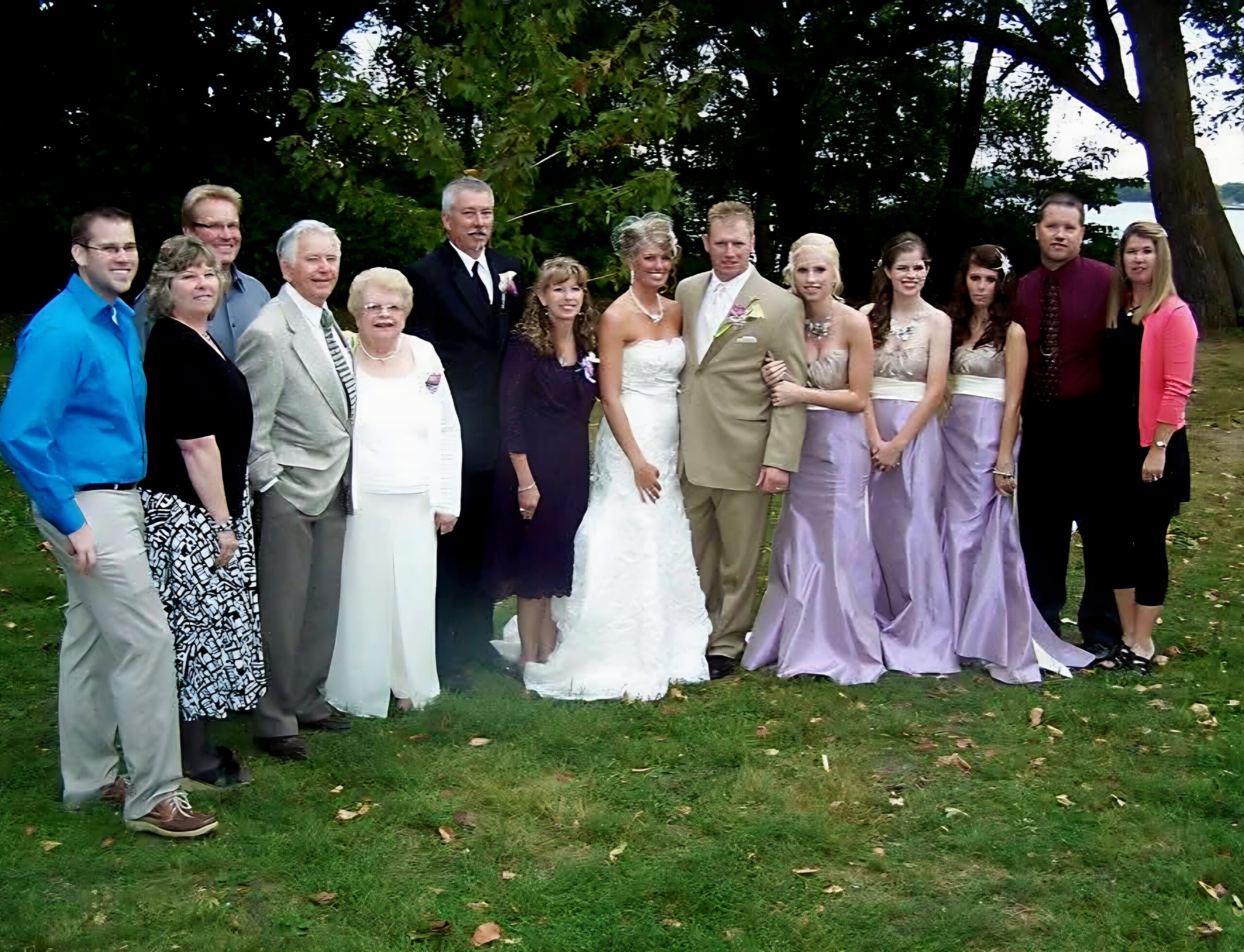 Group of wedding guests posing outdoors with bride and groom in center, bridesmaids in lavender dresses to the right, guests in suits and dresses on a grassy area with trees behind.