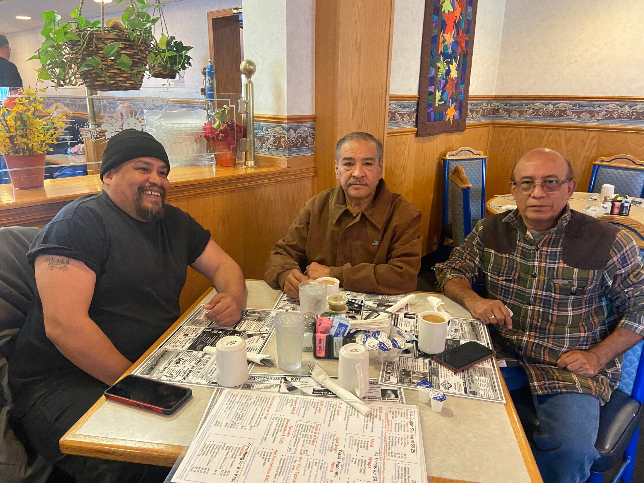 Three men sit at a booth in a diner, drinks on a newspaper-covered table.