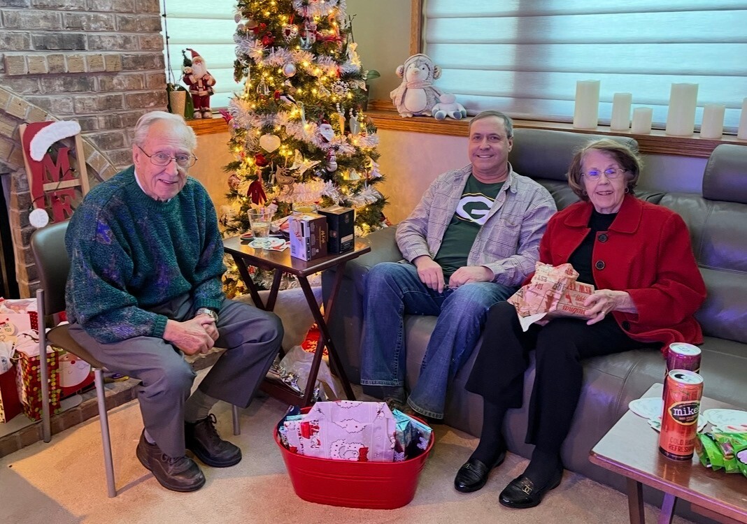 Three adults sit around a lit Christmas tree with wrapped gifts on the floor and a cozy holiday living room scene.