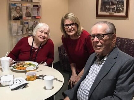 Three seniors sit around a small table, eating and chatting in a cozy dining space; cups and plates are on the table. Best friends or colleagues enjoying a meal together.