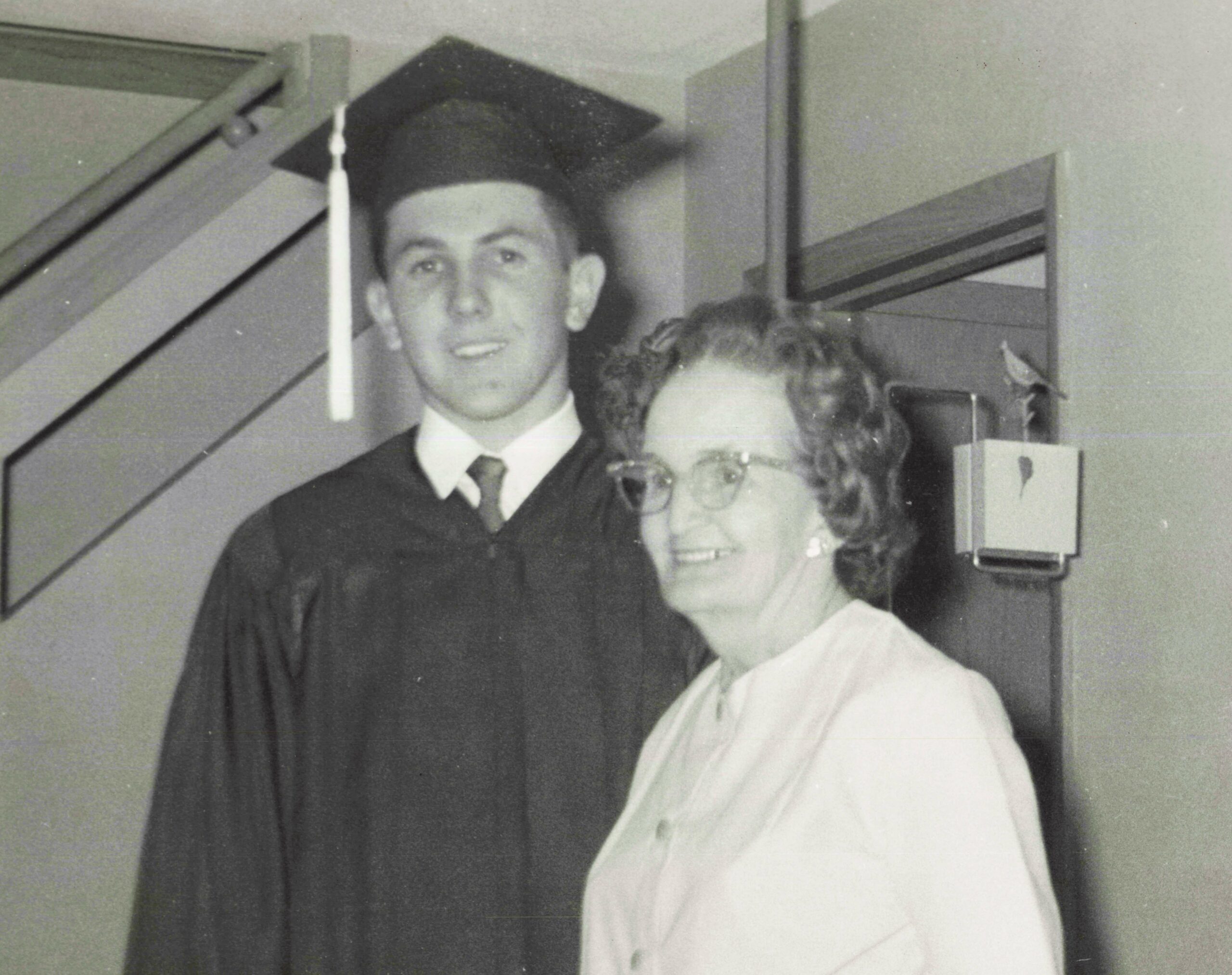 Young man in graduation gown and cap with tassel, standing beside a woman wearing glasses indoors.