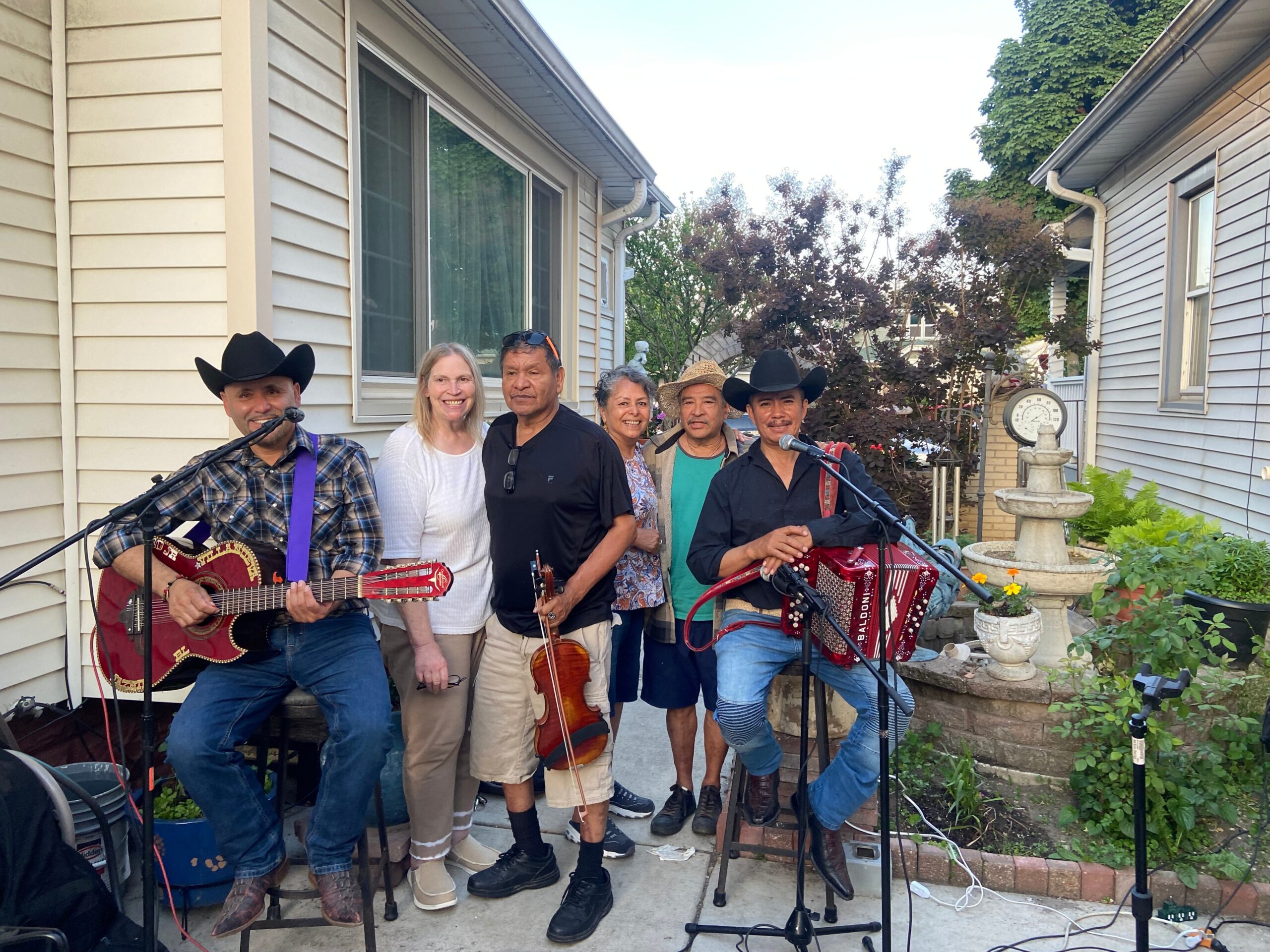 Group of six adults with guitars and microphones posing in a backyard with siding houses in the background.