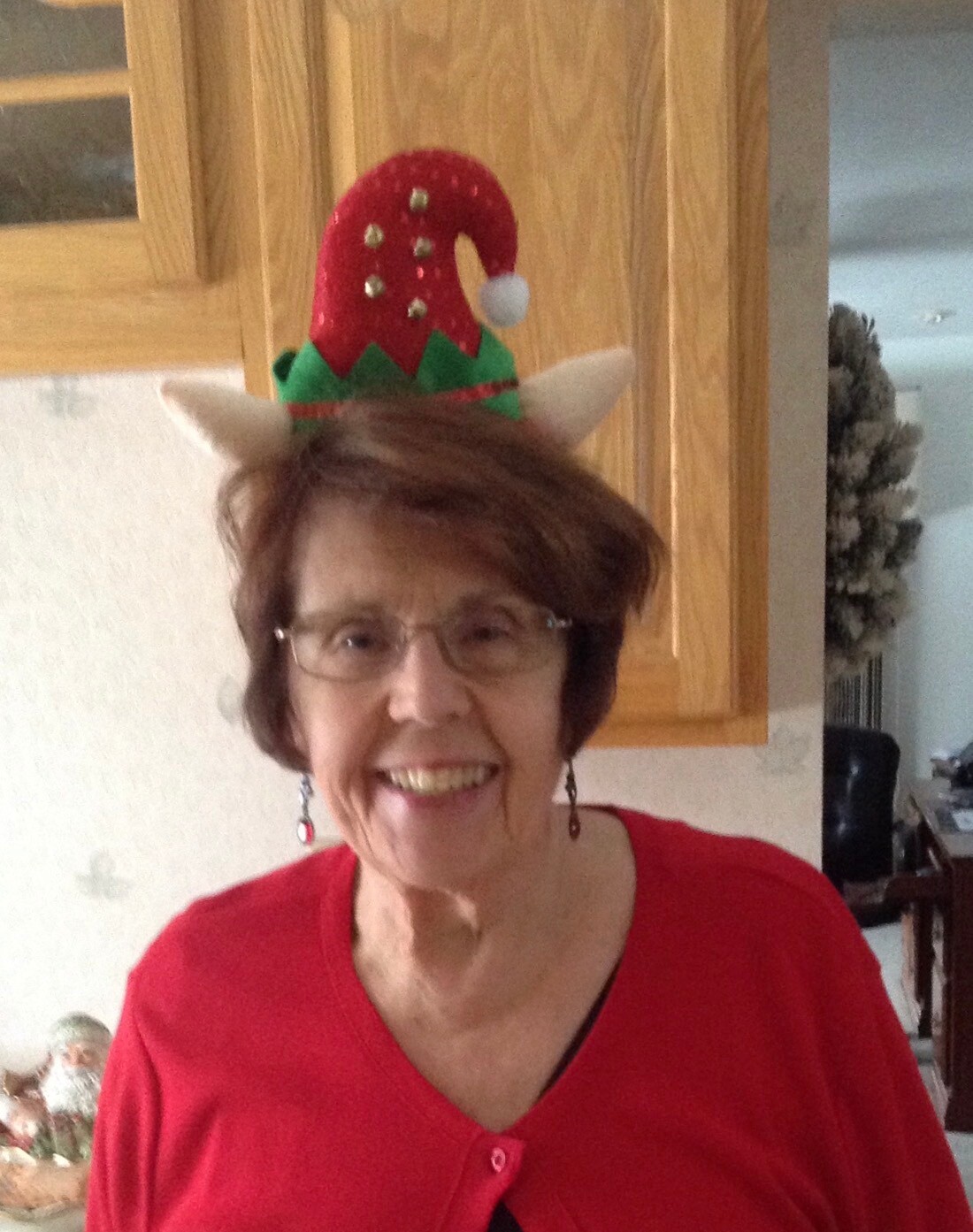 Smiling older woman in a red top wearing a festive elf hat in a kitchen setting.