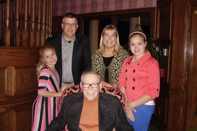 Smiling elderly man in a brown blazer sits in a red-patterned armchair as his family stands behind him in a wood-paneled room, posing for a photo.
