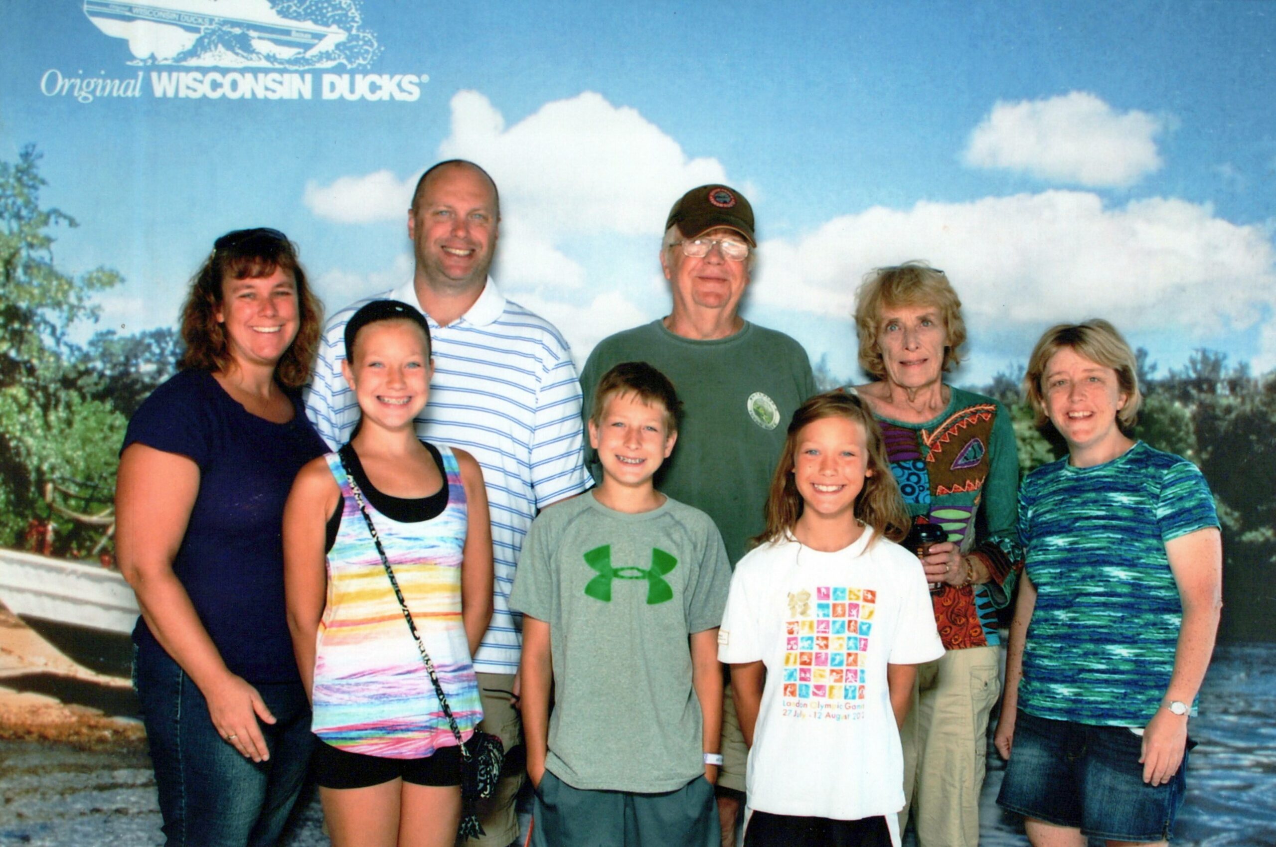 Group of eight people posing for a photo, smiling outdoors in front of a blue sky backdrop with 'Original WISCONSIN DUCKS' text.
