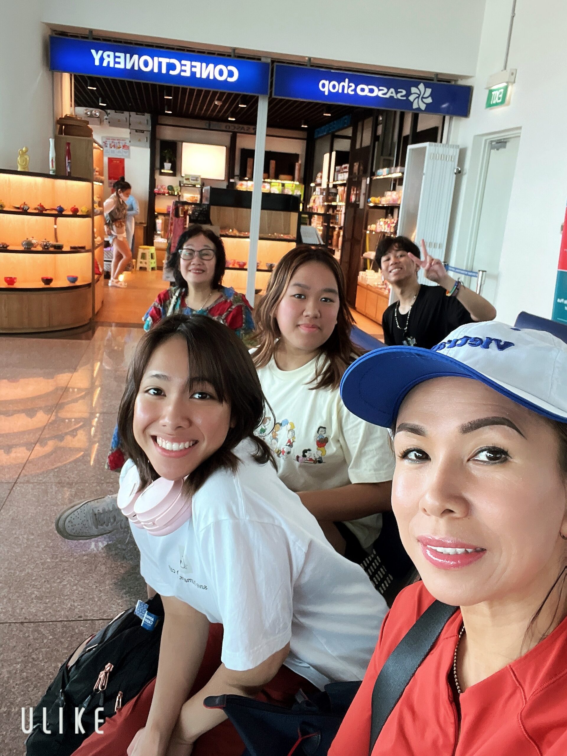 Four friends pose for a selfie in a shopping mall, with store displays and shelves in the background.