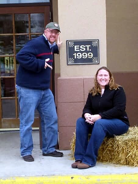 Man in cap and blue sweater makes a gesture beside a doorway; a woman sits on a hay bale smiling, next to a sign that reads EST. 1999 outside a building.