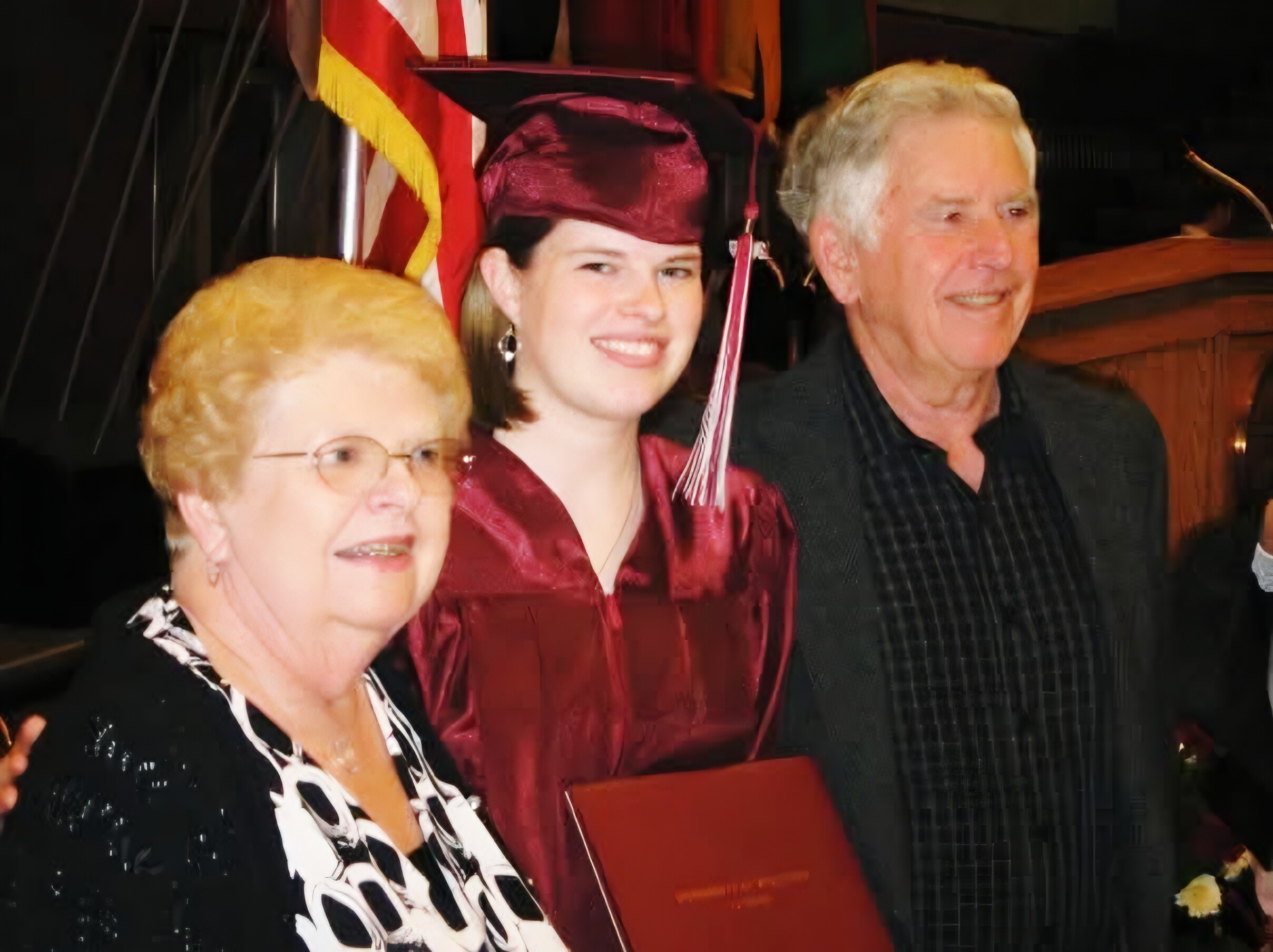 Smiling graduate in a burgundy cap and gown stands with an elderly woman and an older man at night, a flag visible behind them.