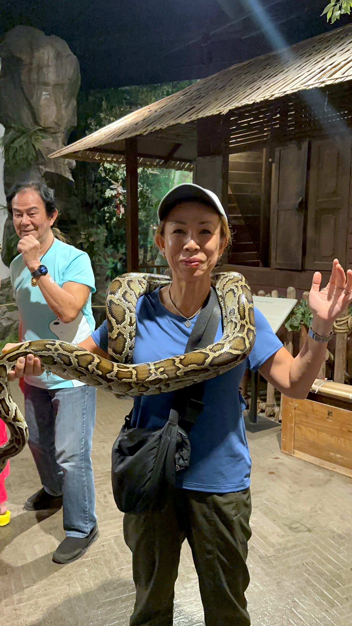 Woman in a blue shirt and cap with a large snake draped around her shoulders indoors at a reptile exhibit.