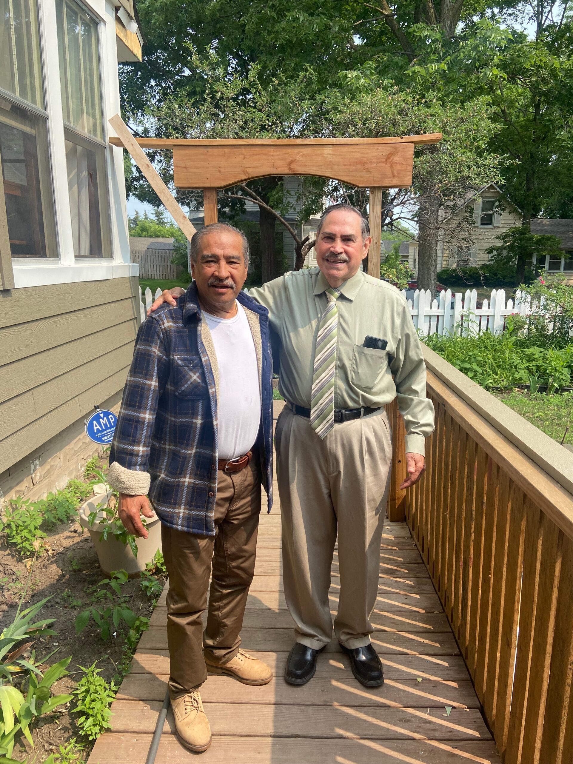Two men pose together on a wooden deck in a residential backyard, with a small wooden frame under construction behind them.