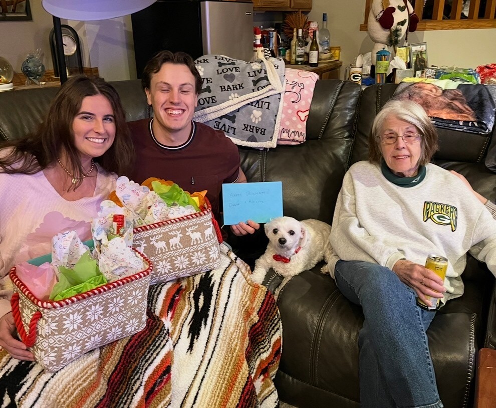 Three people sit on a living room couch with two gift baskets, a small white dog with a red bow, a blue greeting card, and cozy blankets in the background, smiling at a celebration.