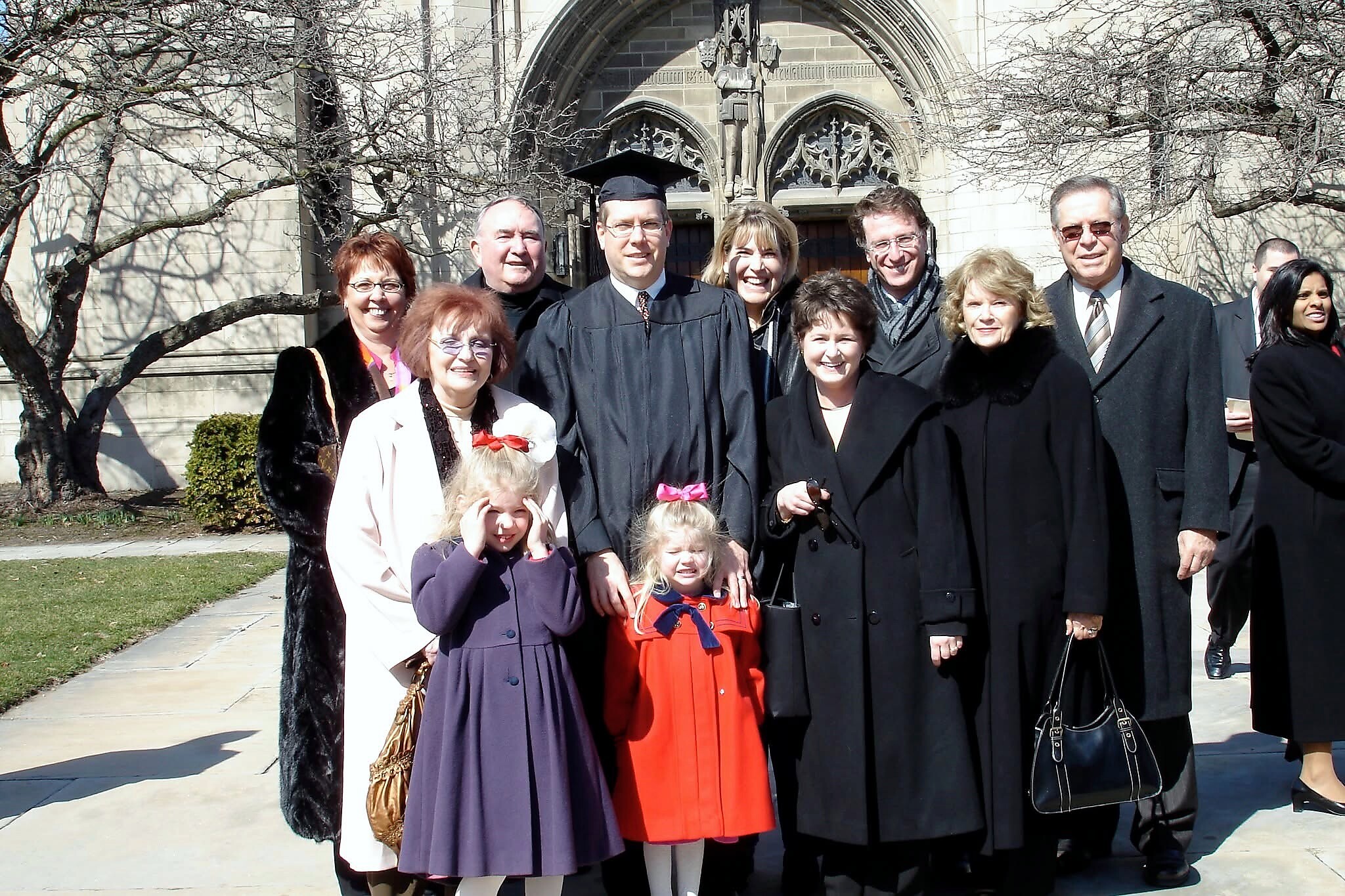 Group of graduates and family posing for a photo outside a stone building, with a graduate in cap and gown front center and children in front.