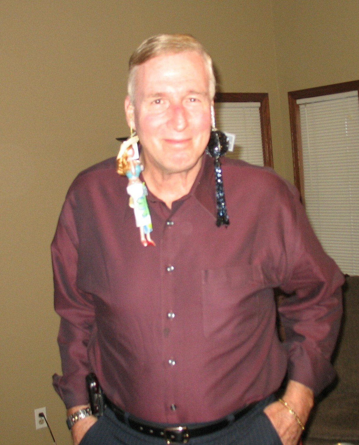 Older man in a maroon dress shirt posing with playful, oversized earrings made of dolls hanging from his left ear